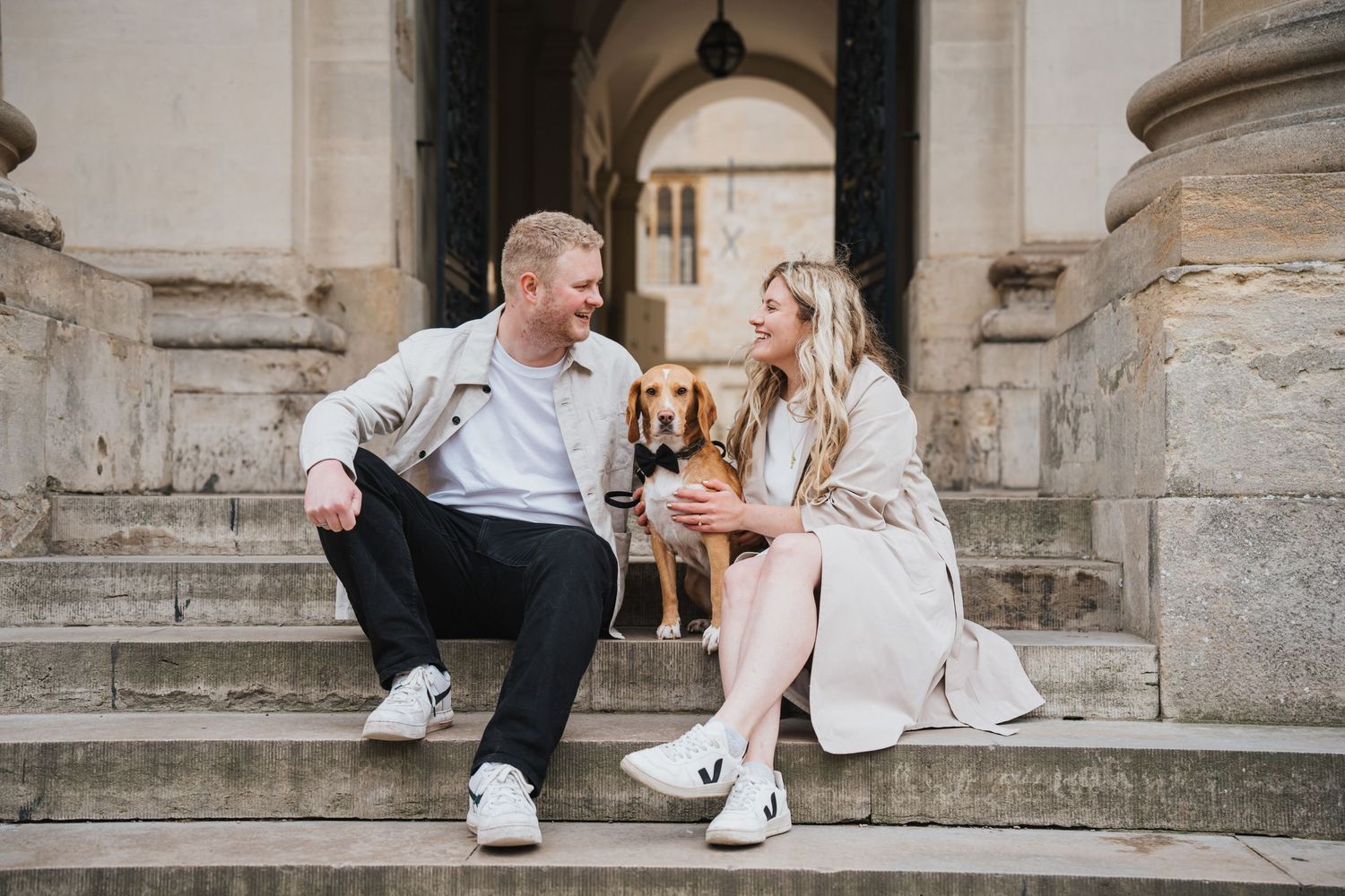 A smiling couple holds hands outdoors near stone columns, dressed in elegant casual attire on a cloudy day.