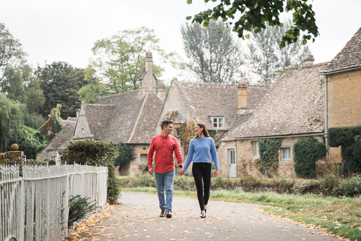 Couple holding hands walking along a charming Cotswolds village path with stone cottages and autumn trees.