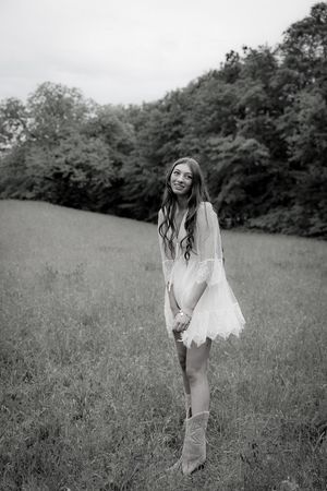 Young woman in white lace dress and cowboy boots standing in a green meadow, black and white photo.