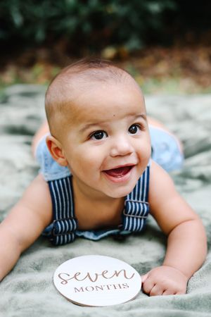 Adorable 7-month-old baby boy smiling while doing tummy time with a milestone marker card outdoors.