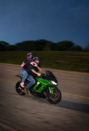 A couple riding together on a green Kawasaki motorcycle at dusk wearing matching pink helmets on a highway.