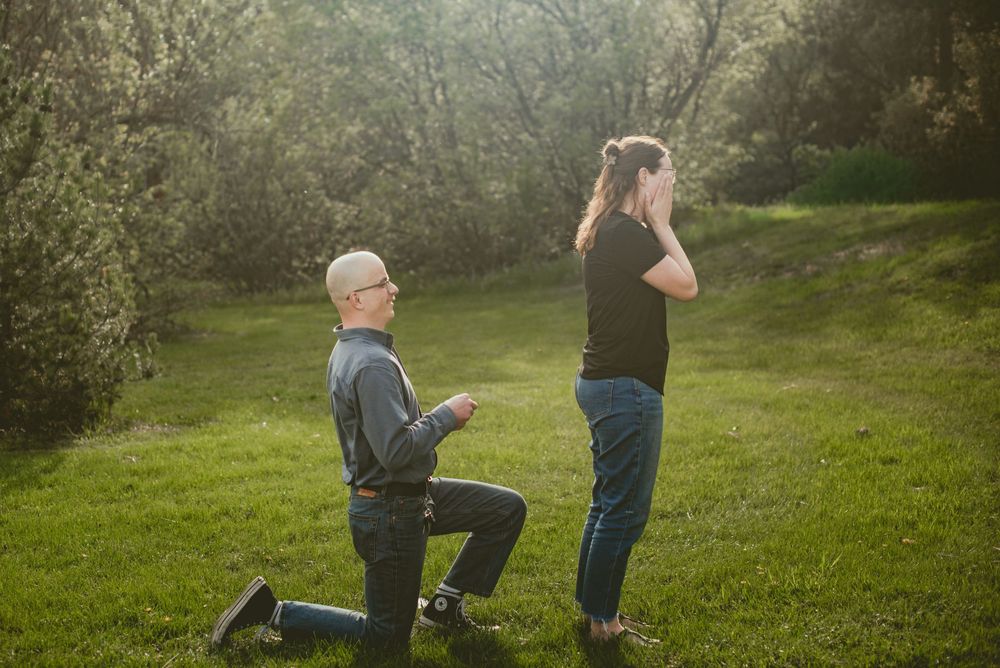 Man kneeling on one knee proposing to woman in lush green park, romantic outdoor marriage proposal scene.