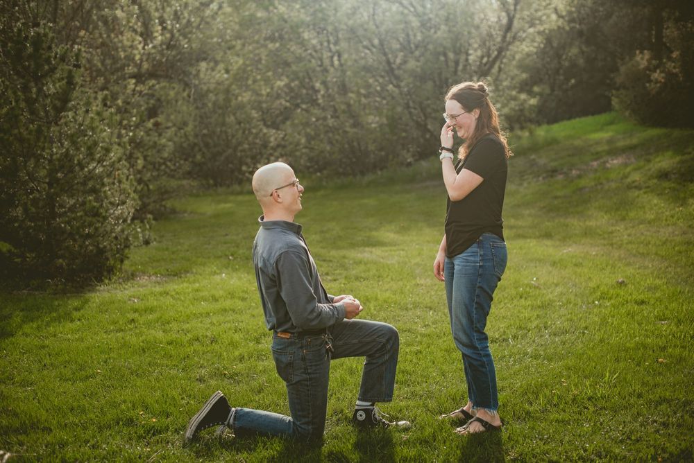 Man proposes on one knee to emotional woman in sunlit green park surrounded by blooming trees.