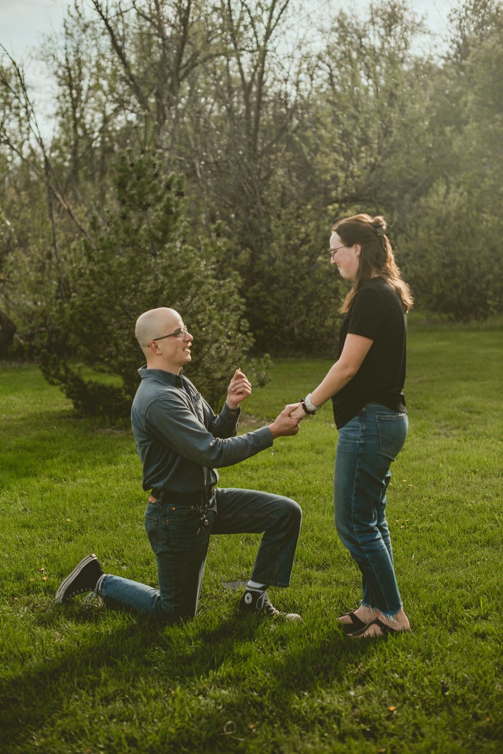 Bald man in glasses proposing on one knee to woman in black top and jeans outdoors.
