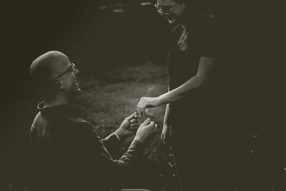 Black and white photo of man on one knee holding womans hand during outdoor proposal.