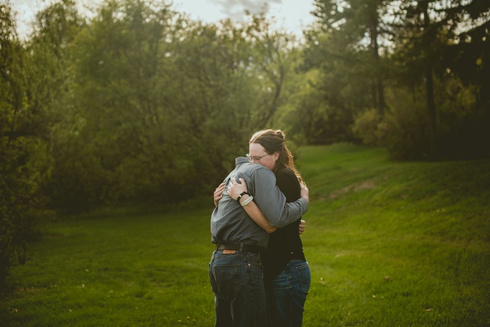 Couple embracing in a warm hug on green grass surrounded by trees after proposal.