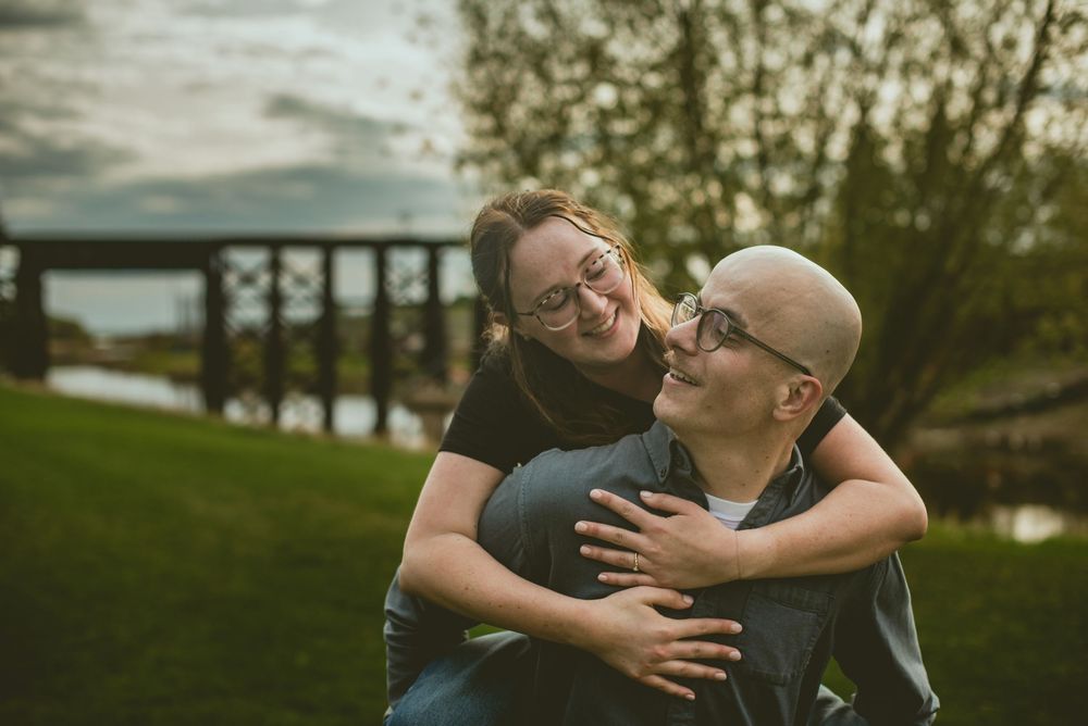 Couple laughing and embracing outdoors in a park with a bridge and river in the background.