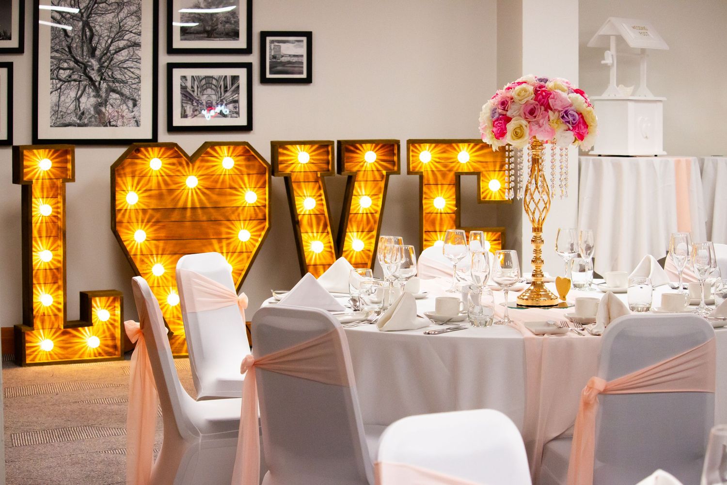 Elegant wedding reception with illuminated LOVE marquee letters, white draped chairs, and pink floral centerpiece.