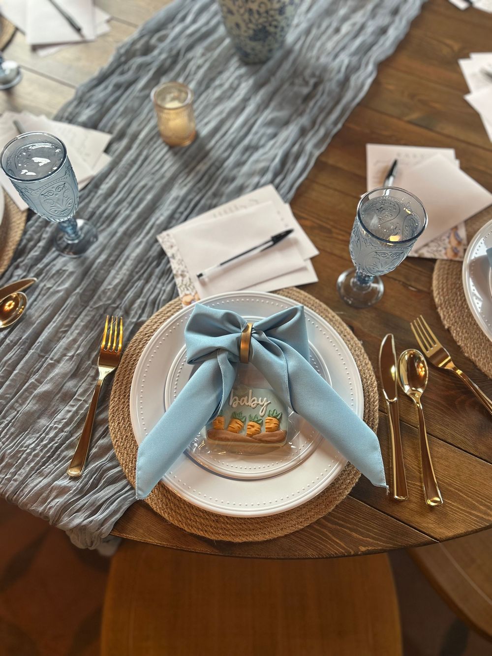 Elegant baby shower table setting with blue bow napkin, gold cutlery, woven charger, and dusty blue glassware on wood table.