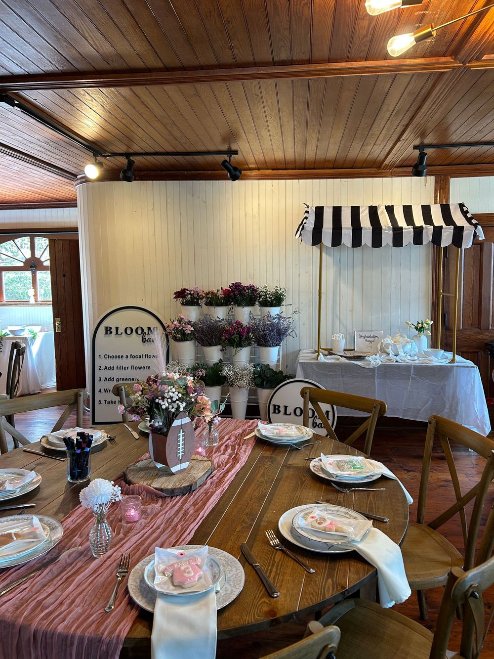 Rustic event venue with floral bar display, striped awning, and elegantly set wooden tables with pink runners.