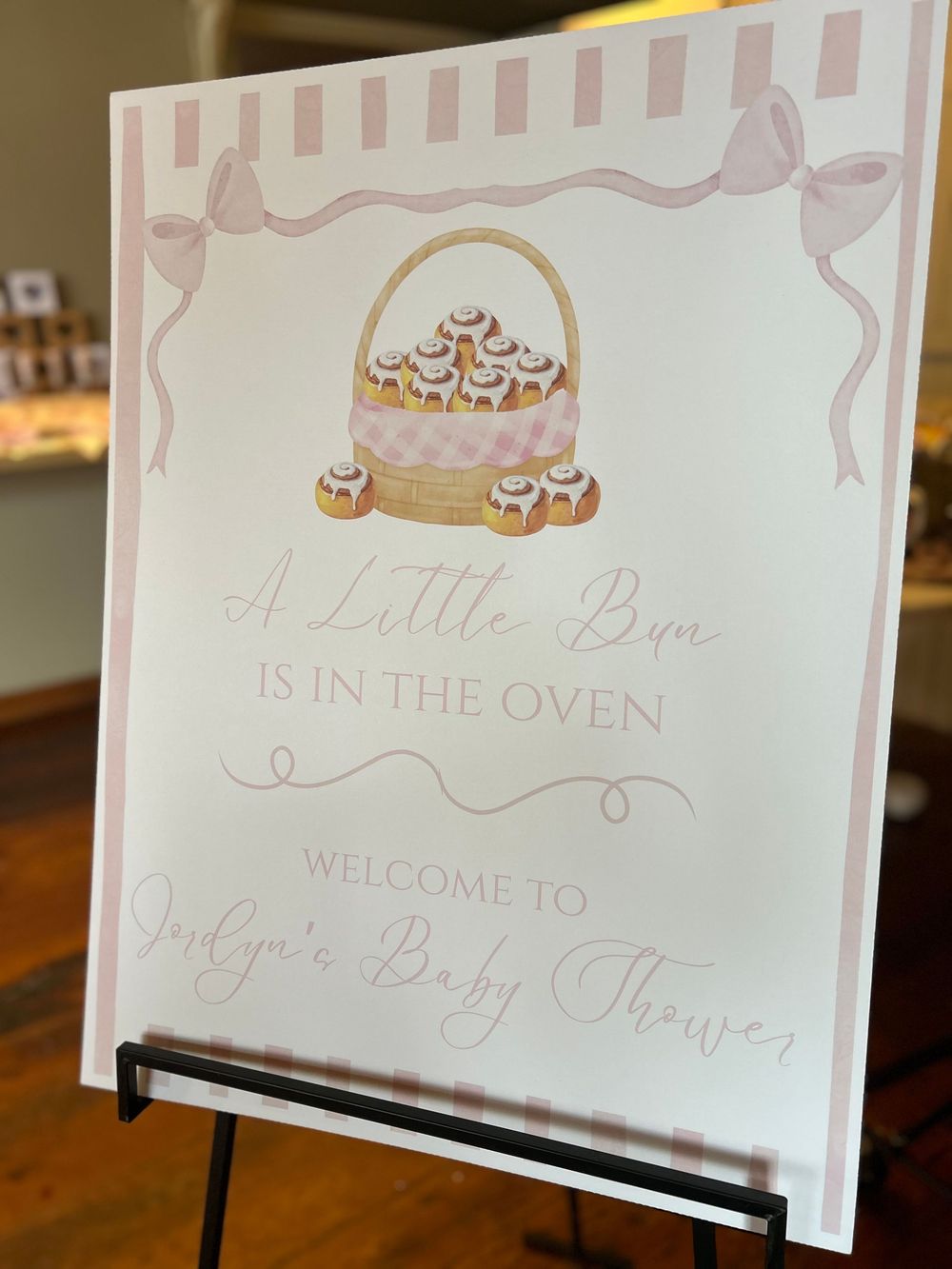 Pink baby shower welcome sign reading A Little Bun Is In The Oven with cinnamon rolls basket illustration.