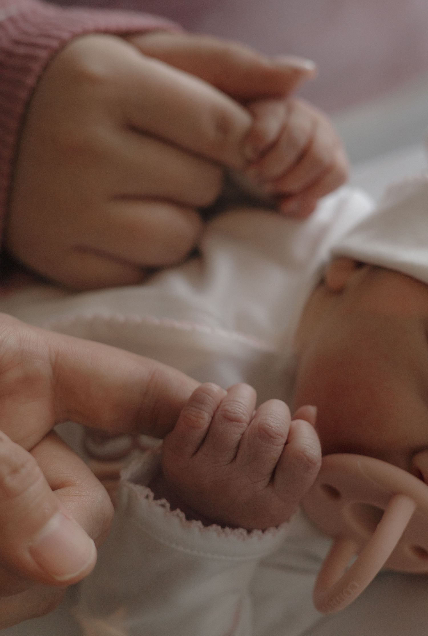Newborn baby sleeping peacefully with a teal pacifier held by an adult hand in a hospital bassinet.
