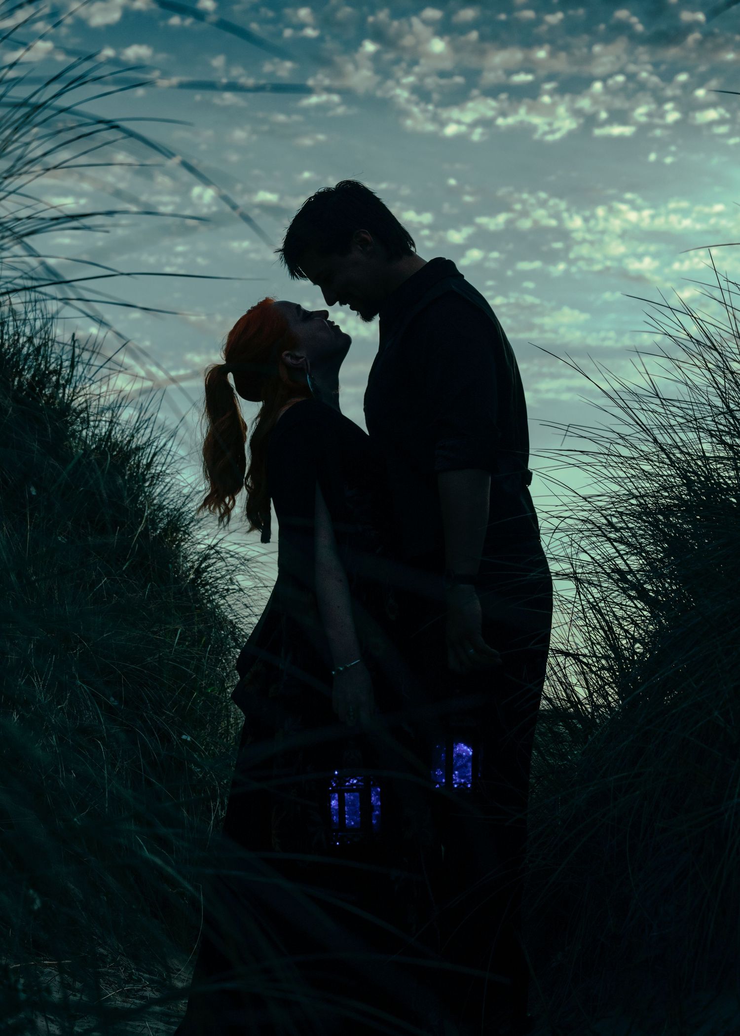 Romantic couple silhouetted against a dramatic twilight sky, sharing an intimate moment among tall beach grasses at dusk.