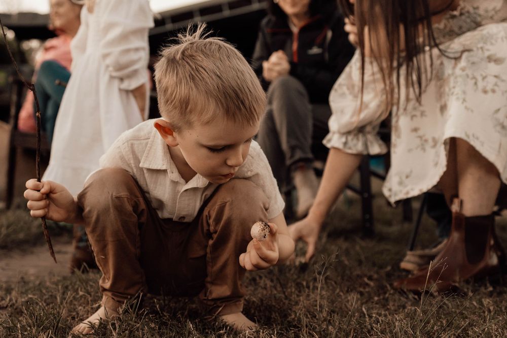 Barefoot boy crouching on grass examining sticks at an outdoor family gathering.