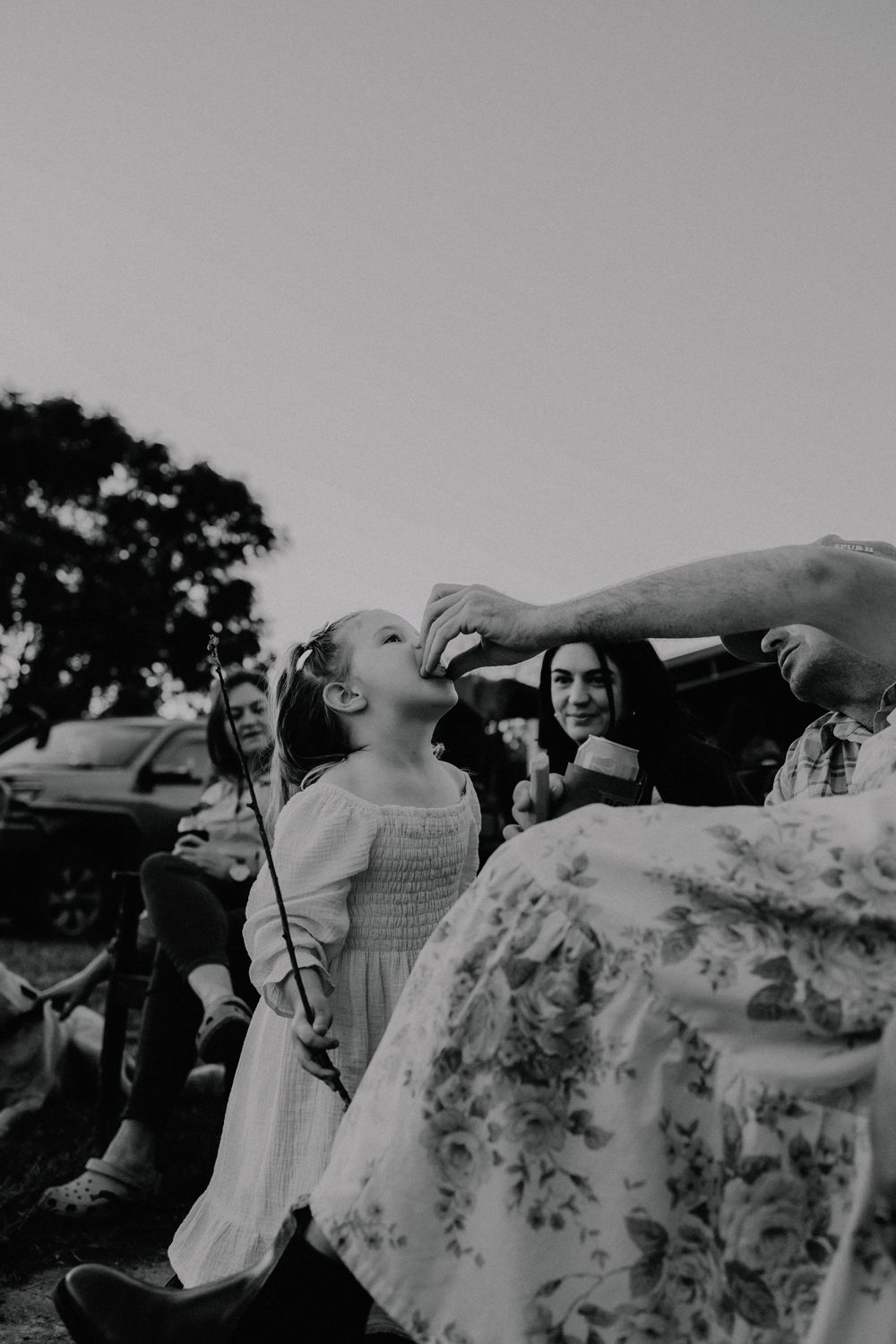 Black and white photo of laughing girl in white dress being fed a treat by an adult at family gathering.