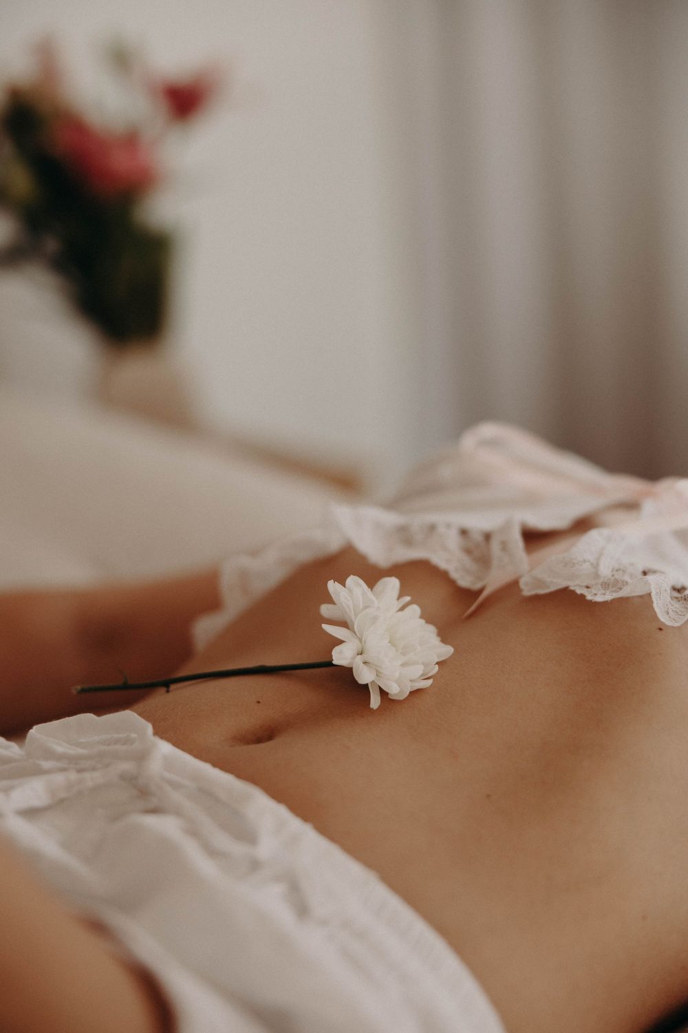 A white floral hair pin resting on a woman's bare midriff, wearing delicate lace lingerie, with soft bokeh background.