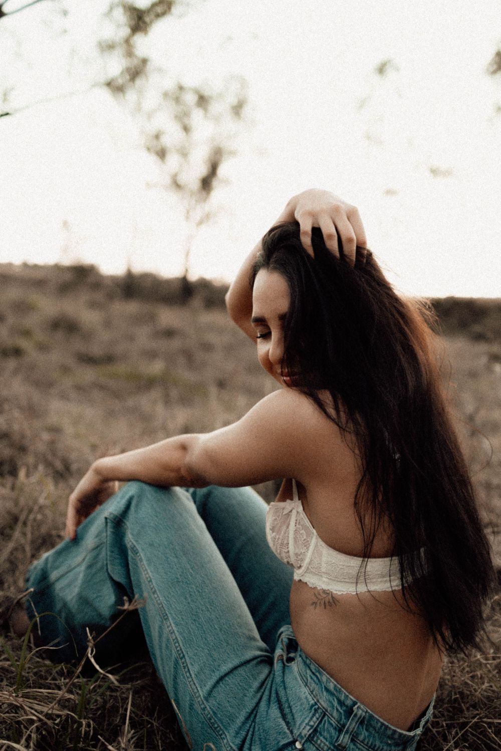 Young woman with long dark hair sits in a field wearing a white bralette and ripped jeans.