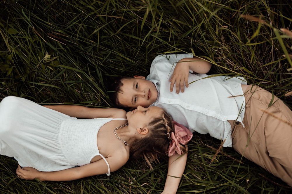 Two children lying in green grass, smiling and embracing in a candid outdoor portrait.