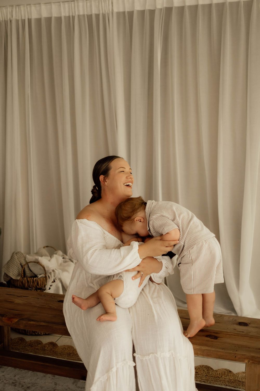 Laughing mother in white dress holding baby while toddler hugs her in a bright, airy room with white curtains.