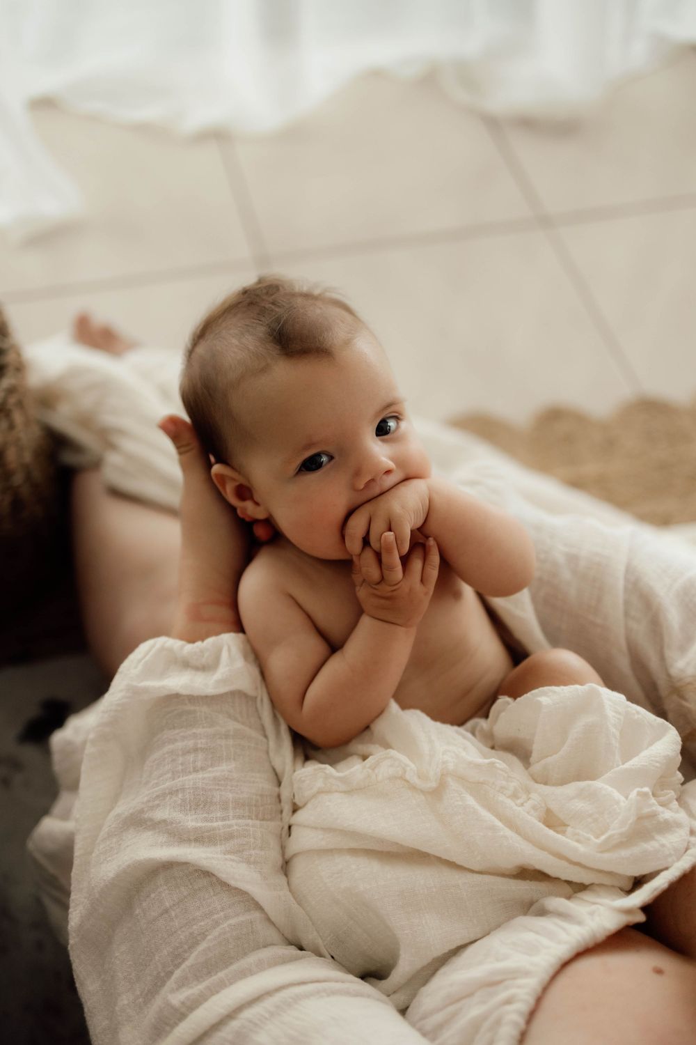 Adorable newborn baby wrapped in white muslin cloth sucking fingers while being held by mother.