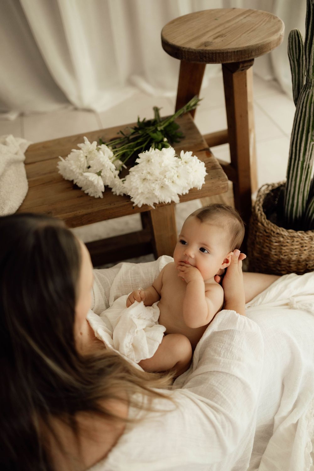 Mother in white dress bonding with curious newborn baby on floor near wooden stool and white flowers.