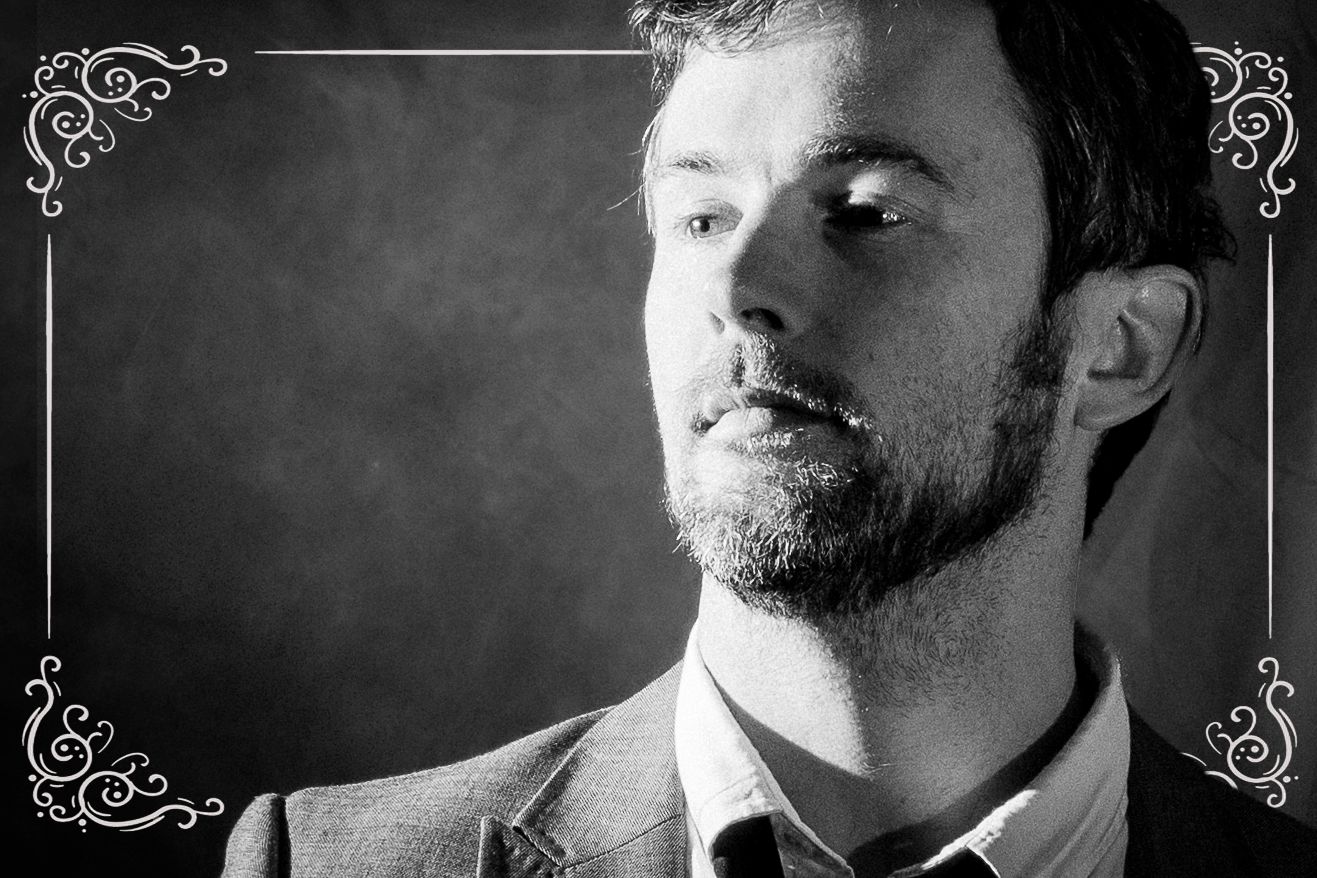 Black and white portrait of a bearded man in a suit jacket looking sideways against a dark background.
