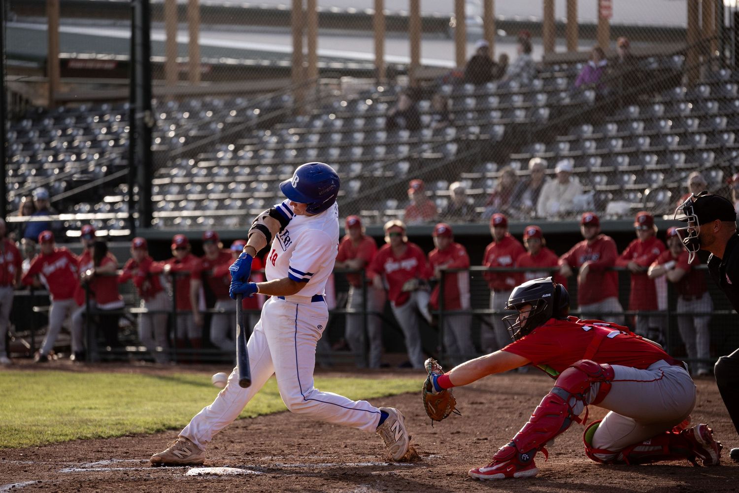 Brooks Bombers (WCBL) batter in blue uniform swings at pitch while catcher crouches behind home plate in stadium.
