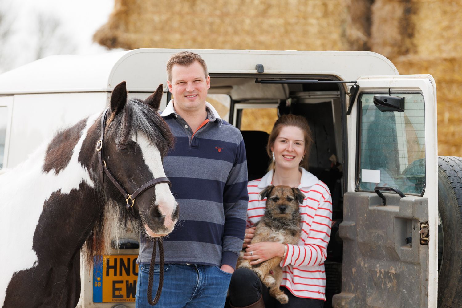 A smiling couple stands with a black and white horse and small dog in front of a white van on a farm.
