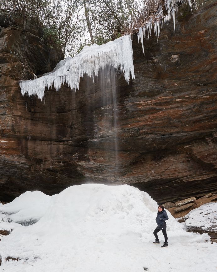 Person standing before a frozen waterfall with icicles on red rock cliffs surrounded by snow in winter.