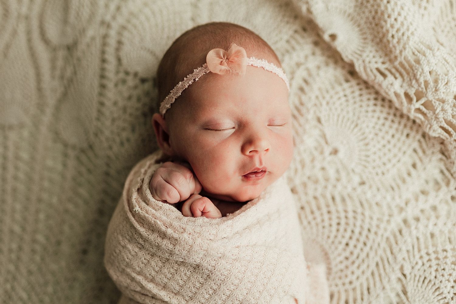 Peaceful newborn baby girl swaddled in cream wrap with floral headband, sleeping on knit blanket.