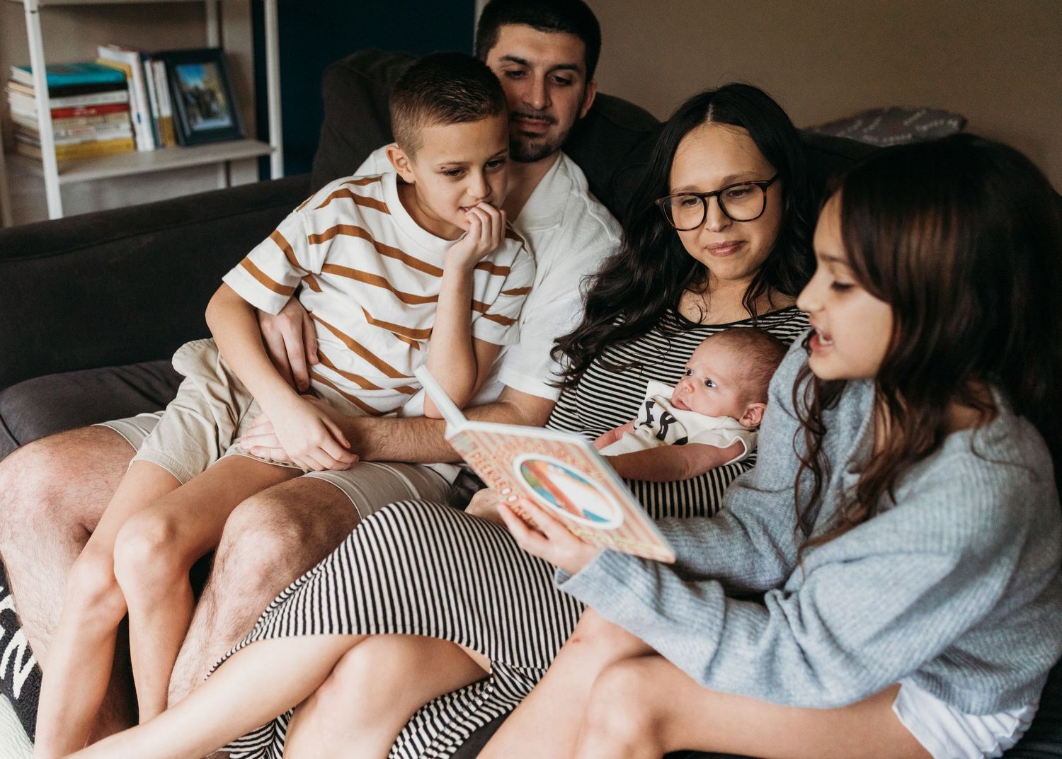 Family of five sitting on couch, parents with three children including newborn baby, sharing a tender moment together.