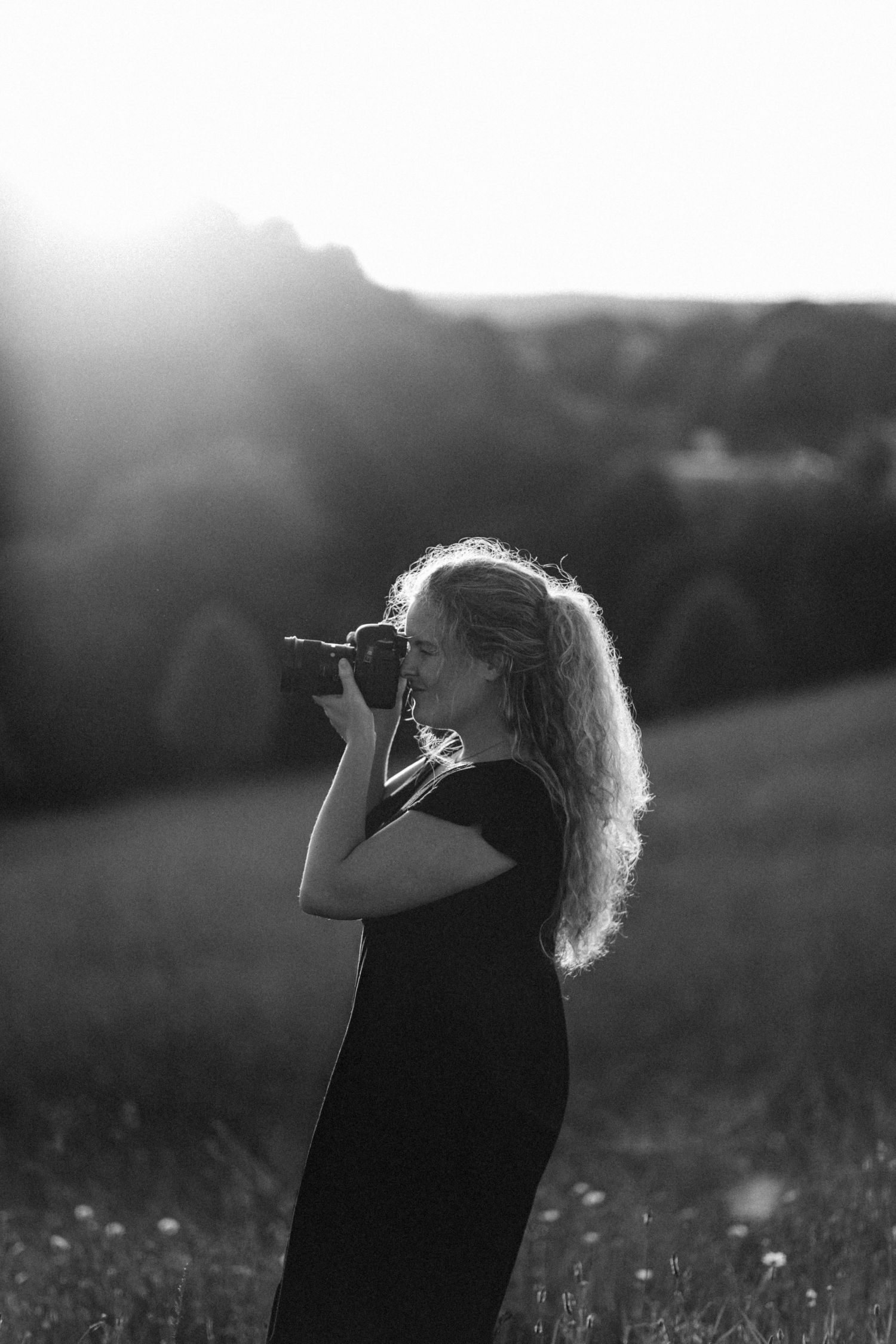 Black and white side profile of a woman taking photos with a camera in a sunlit meadow.