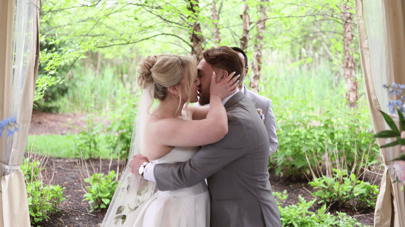 A bride and groom share their first kiss at an outdoor wedding ceremony surrounded by lush green trees and flowers.