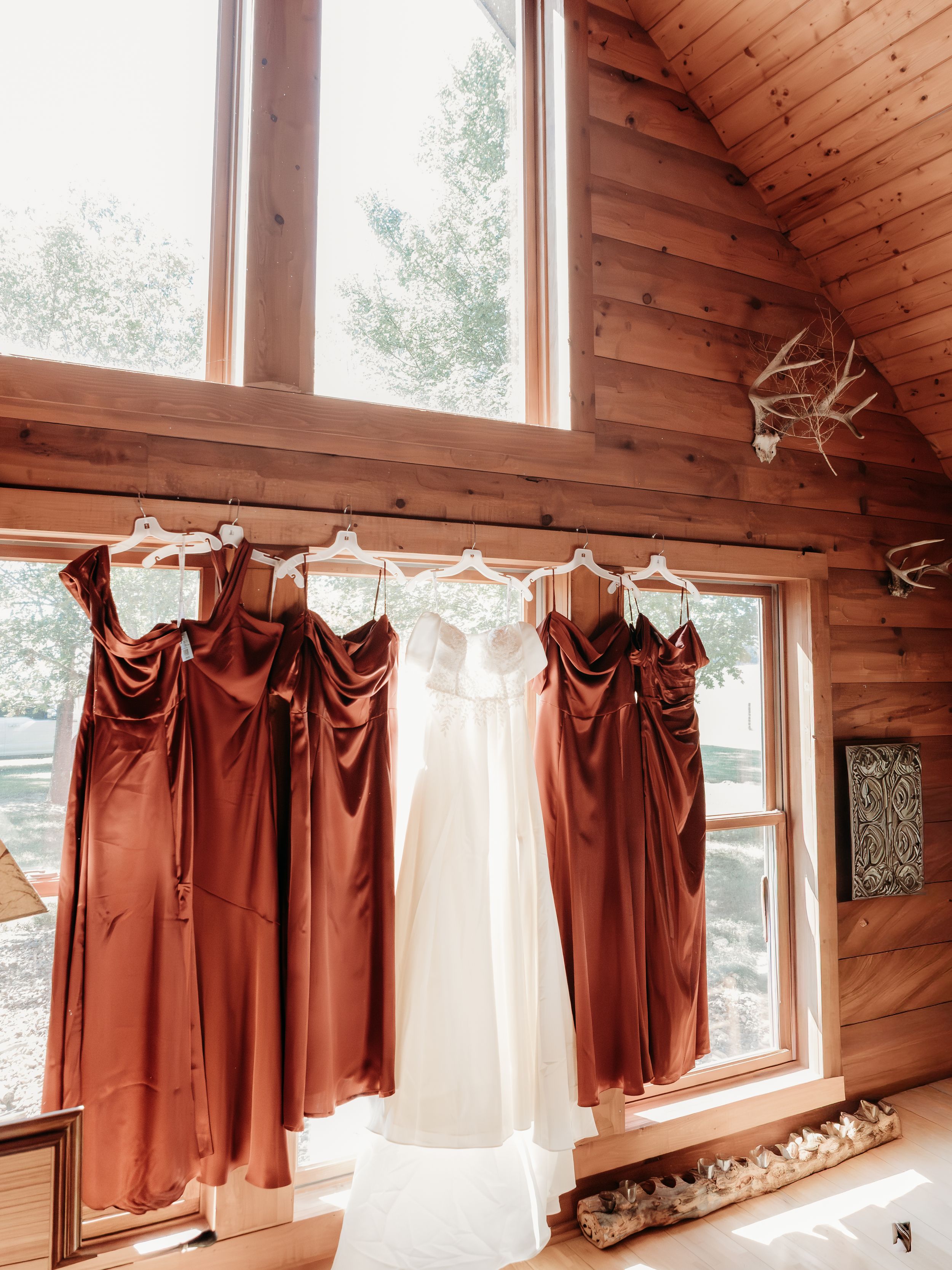 Rust bridesmaid dresses and white bridal gown hanging in a rustic log cabin with large windows.