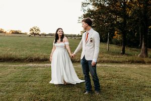 Bride and groom holding hands walking in green field at golden hour, bride in white off-shoulder gown, groom in white blazer.
