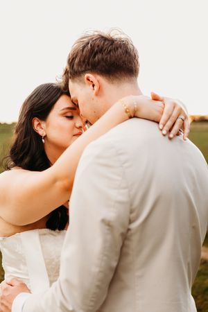 A bride and groom share a tender moment embracing in a golden field at sunset, wearing white wedding attire.