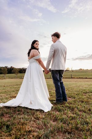 Bride in white off-shoulder gown holds hands with groom in tan blazer and jeans in a scenic outdoor field.