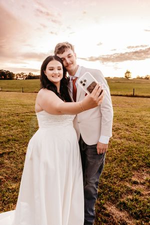 Bride and groom taking a selfie at sunset in a green field, wearing white dress and suit.