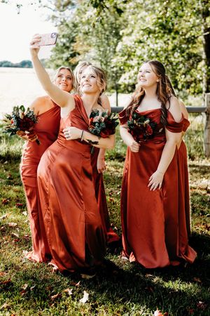 Three bridesmaids in rust satin gowns holding bouquets take a selfie outdoors at a wedding celebration.