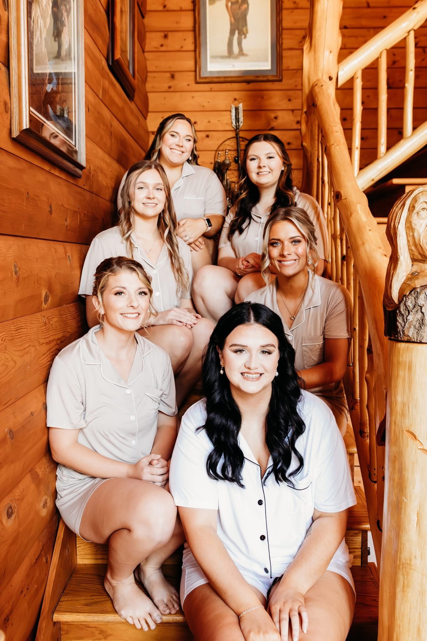Six smiling women in matching pajamas pose together on wooden cabin stairs for a bridal party photo.