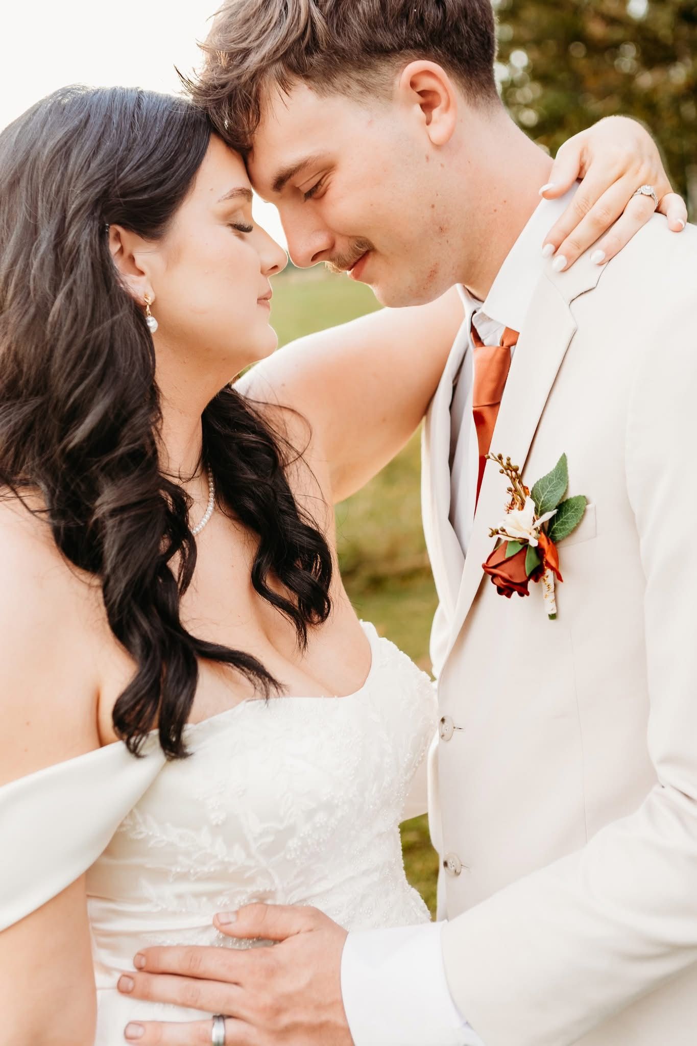 Bride and groom in white dress and cream suit with rose boutonniere share an intimate moment, foreheads touching outdoors.