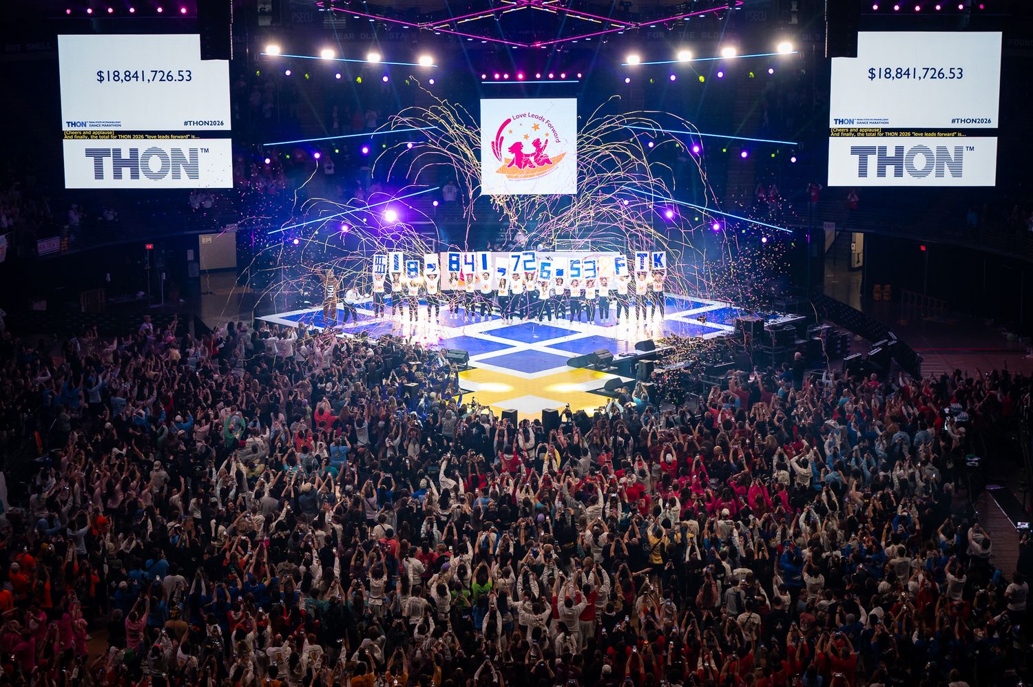 Aerial view of THON fundraiser event at Penn State, packed arena with colorful lights and fundraising total displayed.