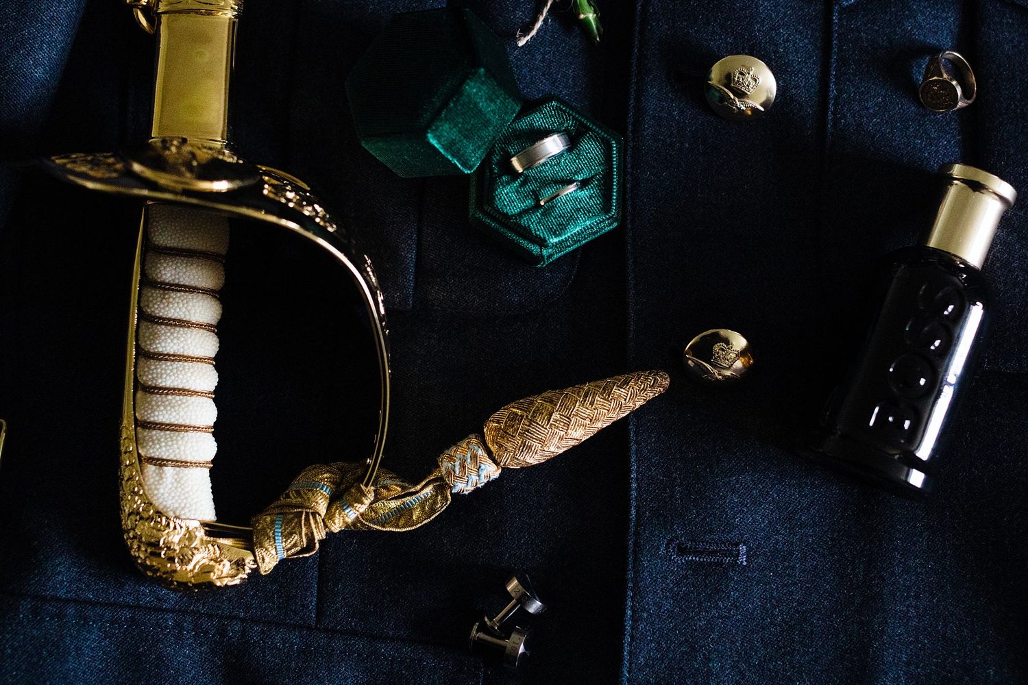 Gold-handled ceremonial sword displayed with rings, cufflinks, and a black perfume bottle on a dark navy suit.
