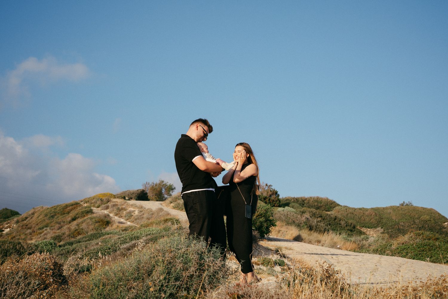 Couple sharing a romantic moment outdoors on a sunny hillside, dressed in black casual wear against a blue sky.