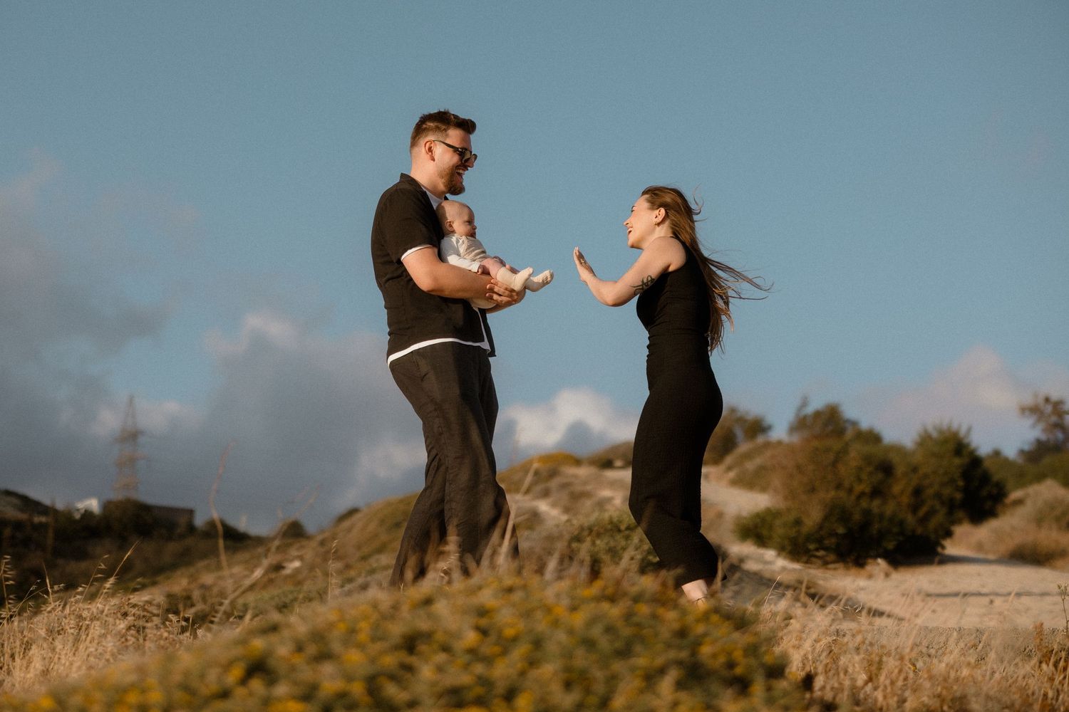 Couple sharing a romantic moment outdoors on a sunny hillside, dressed in black casual wear against a blue sky.