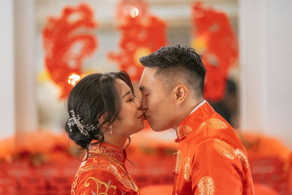 Couple sharing a kiss in traditional red Chinese wedding attire with decorative double happiness symbols in background.