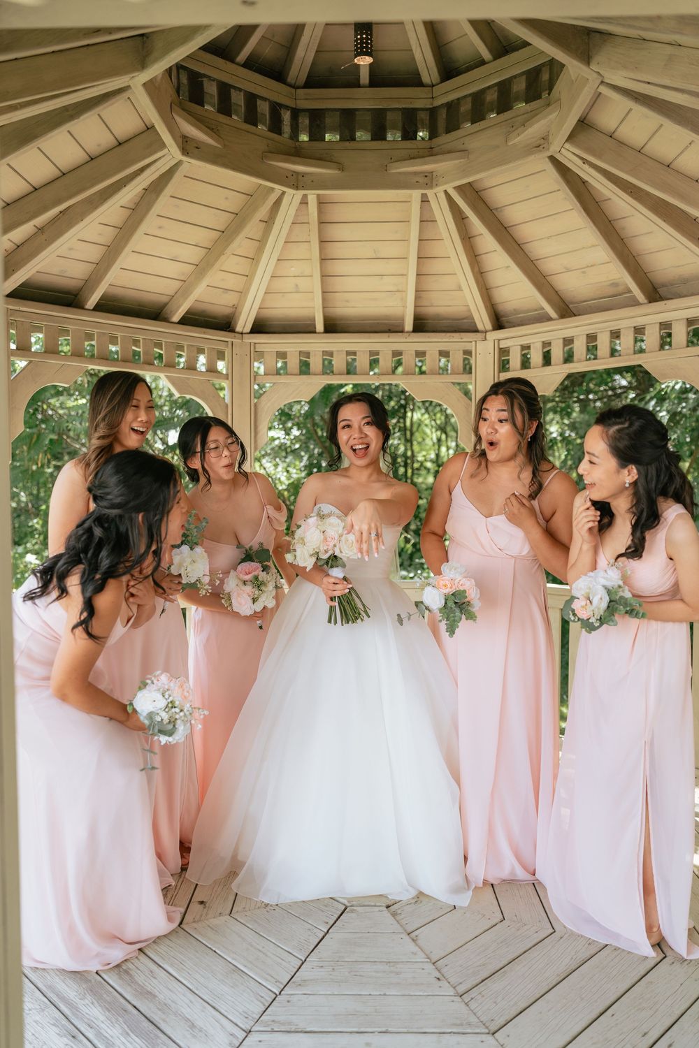 Bride laughing with bridesmaids in pink gowns holding bouquets inside a wooden gazebo.