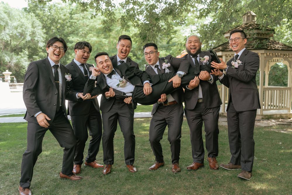 Groomsmen in dark suits lifting the groom outdoors near a gazebo, celebrating at a wedding party.