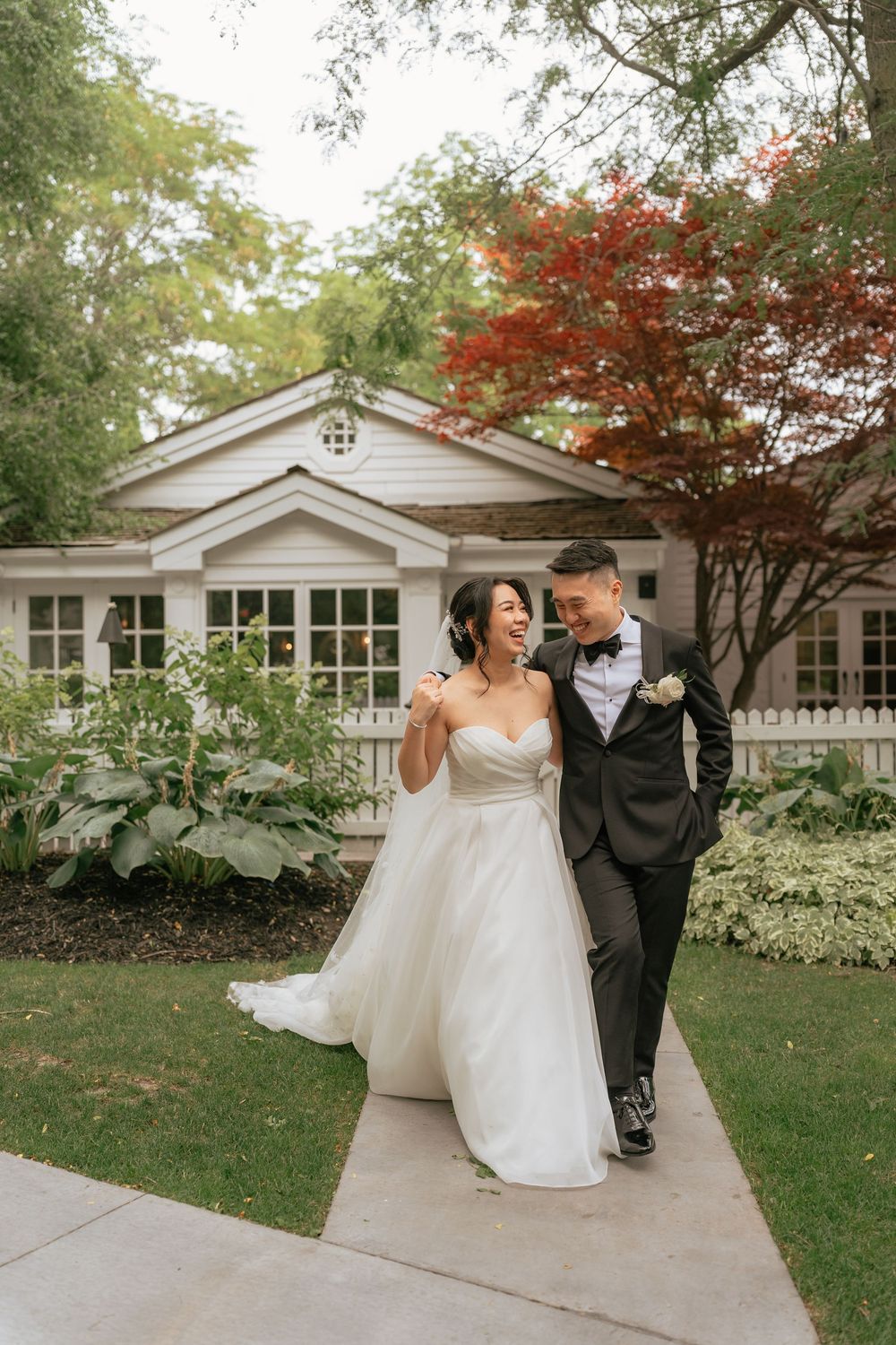 Bride in white gown and groom in black tuxedo walking and laughing outside a charming white cottage.