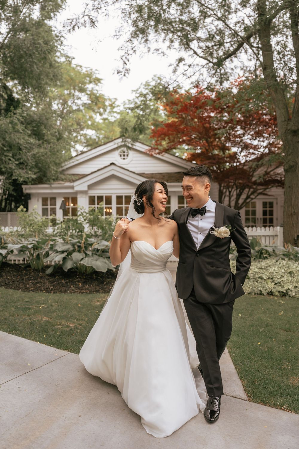 Smiling Asian bride and groom walk together in wedding attire outside a charming white cottage surrounded by lush greenery.